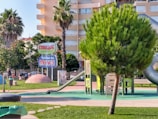 A colorful playground with several slides and climbing structures surrounded by greenery. The area is well-maintained with vibrant grass and a few palm trees. A prominent sign displays the name 'Parque Turma da Mônica'. Several buildings can be seen in the background.