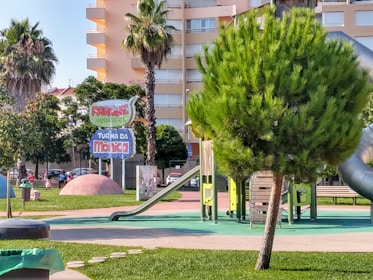 A colorful playground with several slides and climbing structures surrounded by greenery. The area is well-maintained with vibrant grass and a few palm trees. A prominent sign displays the name 'Parque Turma da Mônica'. Several buildings can be seen in the background.