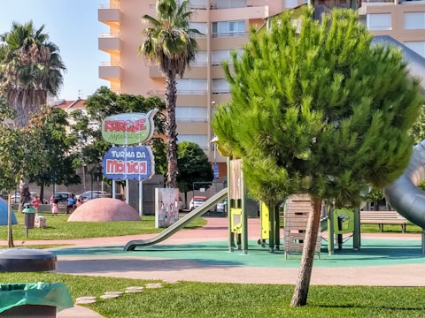 A colorful playground with several slides and climbing structures surrounded by greenery. The area is well-maintained with vibrant grass and a few palm trees. A prominent sign displays the name 'Parque Turma da Mônica'. Several buildings can be seen in the background.