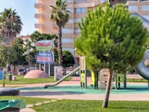 A colorful playground with several slides and climbing structures surrounded by greenery. The area is well-maintained with vibrant grass and a few palm trees. A prominent sign displays the name 'Parque Turma da M&ocirc;nica'. Several buildings can be seen in the background.