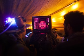 A person has a TV set on their head, creating a surreal and intriguing scene. They are surrounded by other people in a dimly lit environment with vibrant and colorful lighting. The setting suggests a festive or artistic event.