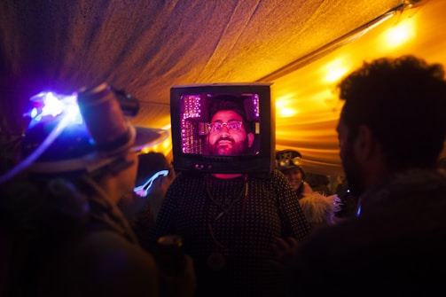 A person has a TV set on their head, creating a surreal and intriguing scene. They are surrounded by other people in a dimly lit environment with vibrant and colorful lighting. The setting suggests a festive or artistic event.