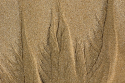 Fine patterns and lines in the sand create an intricate and natural texture. The surface appears soft yet slightly grainy, with gentle shadows highlighting the grooves and valleys formed by the natural movements of wind or water.