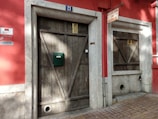 A rustic red building facade featuring two wooden doors with distinctive diagonal planks. The number 21 is displayed above one door, which also has a green mailbox affixed. A sign above reads 'Barbearia da Mina.' The surface is accented with stone frames around the doors, and there are circular vents at the base.