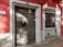 A rustic red building facade featuring two wooden doors with distinctive diagonal planks. The number 21 is displayed above one door, which also has a green mailbox affixed. A sign above reads 'Barbearia da Mina.' The surface is accented with stone frames around the doors, and there are circular vents at the base.