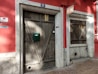 A rustic red building facade featuring two wooden doors with distinctive diagonal planks. The number 21 is displayed above one door, which also has a green mailbox affixed. A sign above reads 'Barbearia da Mina.' The surface is accented with stone frames around the doors, and there are circular vents at the base.