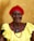 An elderly woman with a warm smile is wearing a bright red headscarf, pearl earrings, and a yellow dress. She has a vibrant red beaded necklace and a blue pendant, standing against a plain beige background.