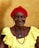An elderly woman with a warm smile is wearing a bright red headscarf, pearl earrings, and a yellow dress. She has a vibrant red beaded necklace and a blue pendant, standing against a plain beige background.
