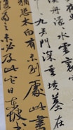 Elegant calligraphy is displayed on textured paper with vertical and diagonal orientations. The script appears in bold black ink against creamy beige and muted gold backgrounds, creating a contrast that highlights the artistry and fluidity of the characters.