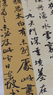 Elegant calligraphy is displayed on textured paper with vertical and diagonal orientations. The script appears in bold black ink against creamy beige and muted gold backgrounds, creating a contrast that highlights the artistry and fluidity of the characters.