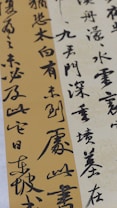 Elegant calligraphy is displayed on textured paper with vertical and diagonal orientations. The script appears in bold black ink against creamy beige and muted gold backgrounds, creating a contrast that highlights the artistry and fluidity of the characters.