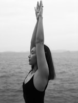 A woman is practicing yoga by the sea, standing with arms raised and eyes closed, conveying a sense of calmness and introspection.