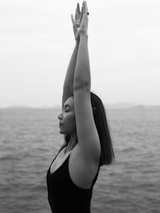 A woman is practicing yoga by the sea, standing with arms raised and eyes closed, conveying a sense of calmness and introspection.