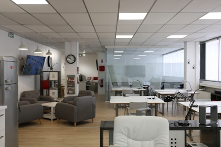 An open office space with modern furnishings, including several tables and chairs, a lounge area with gray sofas, a wall-mounted television, and a refrigerator. The ceiling features recessed lighting, and there are large windows along one side of the room.
