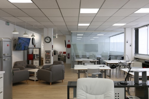 An open office space with modern furnishings, including several tables and chairs, a lounge area with gray sofas, a wall-mounted television, and a refrigerator. The ceiling features recessed lighting, and there are large windows along one side of the room.