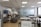 An open office space with modern furnishings, including several tables and chairs, a lounge area with gray sofas, a wall-mounted television, and a refrigerator. The ceiling features recessed lighting, and there are large windows along one side of the room.