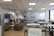 An open office space with modern furnishings, including several tables and chairs, a lounge area with gray sofas, a wall-mounted television, and a refrigerator. The ceiling features recessed lighting, and there are large windows along one side of the room.