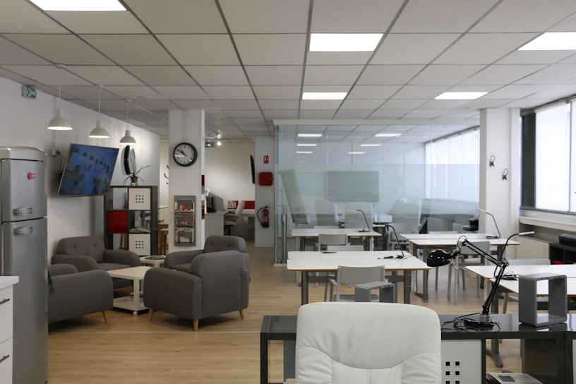 An open office space with modern furnishings, including several tables and chairs, a lounge area with gray sofas, a wall-mounted television, and a refrigerator. The ceiling features recessed lighting, and there are large windows along one side of the room.