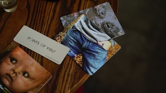 A wooden table features three cards spread out. One card shows an image of a child wearing jeans and a gray shirt, with hands in pockets. Another card displays an old, weathered doll face. The third card, placed diagonally, has the text 'POINTS OF YOU' printed on it. The colors are warm and subdued, with a focus on browns and grays.