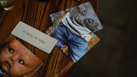 A wooden table features three cards spread out. One card shows an image of a child wearing jeans and a gray shirt, with hands in pockets. Another card displays an old, weathered doll face. The third card, placed diagonally, has the text 'POINTS OF YOU' printed on it. The colors are warm and subdued, with a focus on browns and grays.