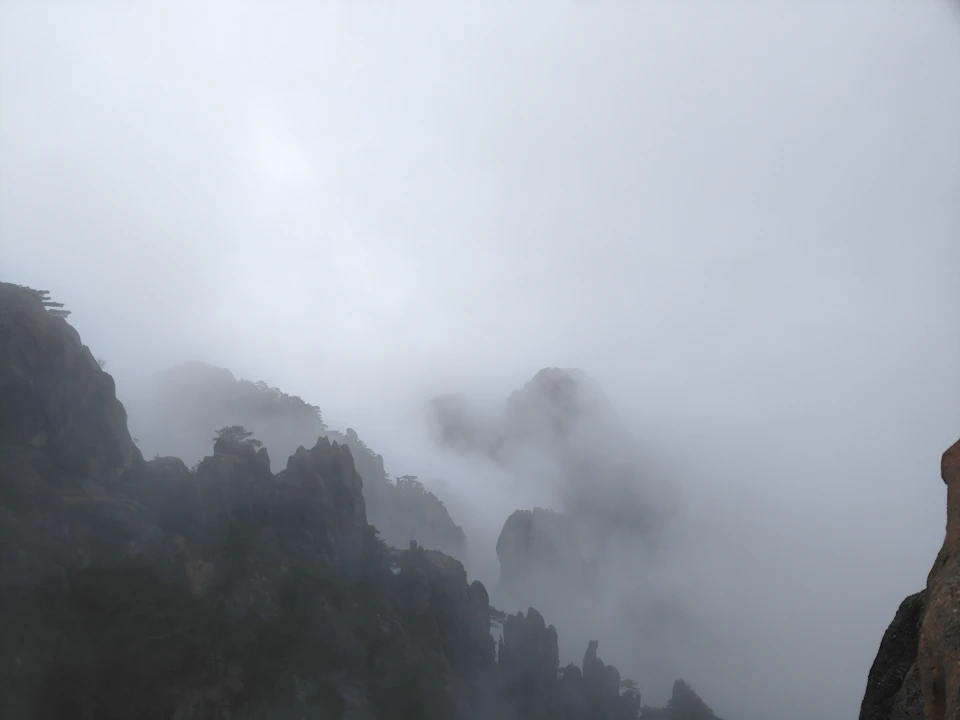 The image depicts a foggy mountain landscape with a large, dark gray cliff on the right side and a smaller, lighter gray cliff on the left side. The fog obscures the view of the mountains, creating an ethereal atmosphere. In the distance, there are trees visible against the foggy backdrop, adding depth to the scene.