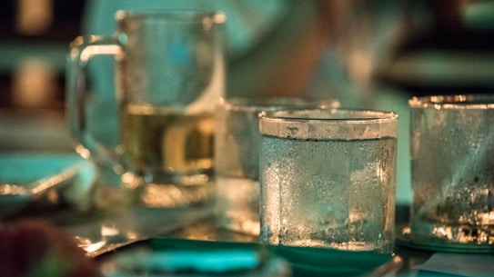 A close-up shot of several frosted glass cups on a table, some filled with beverages. The background is softly blurred, suggesting a dimly lit setting, possibly a bar or a restaurant.