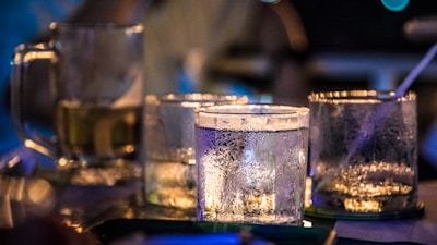 Several glass tumblers and a mug filled with drinks are placed on a table. The glasses are frosty with condensation and the background is blurred, giving a bokeh effect. Low lighting and warm tones create a cozy atmosphere.