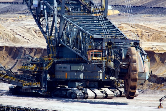 A massive industrial machine used for mining operations, featuring large treads for movement and a giant rotating wheel for excavation. The background shows an open-pit mining environment with exposed rock and soil.