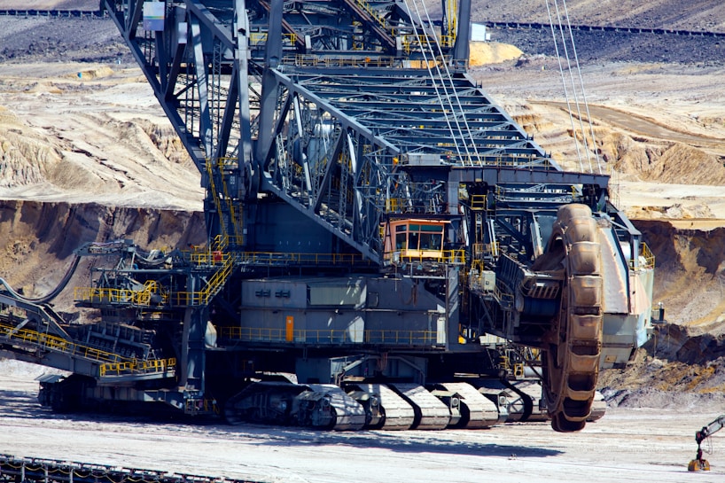 A massive industrial machine used for mining operations, featuring large treads for movement and a giant rotating wheel for excavation. The background shows an open-pit mining environment with exposed rock and soil.