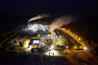 An industrial complex is illuminated at night, emitting bright lights and smoke stacks releasing visible plumes into the dark sky. Structures and machinery are arranged across a large area, with surrounding roads and pathways. The glow of orange-yellow lights highlights the industrial activity.