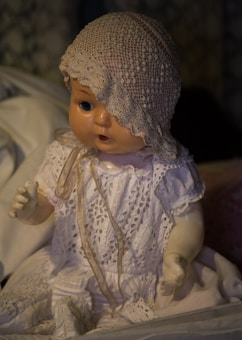 A vintage doll with a lace bonnet and intricate crocheted dress sits in a dimly lit environment. The doll has a slightly open mouth and detailed facial features, creating a somewhat eerie atmosphere. Shadows play across its face, adding to the overall aged and mysterious look.