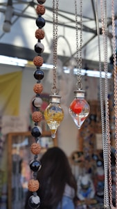 Two necklaces with glass pendants, one filled with yellow liquid and the other with a red object, are displayed. Adjacent is a beaded necklace with alternating brown, black, and cream beads. The background includes chains, a blurred figure, and various market items.