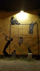 A mural on a yellow wall illuminated by a spotlight features multiple figures, including a person seated above a rectangular frame as if reading, a child on a swing within the frame, a person playing a wind instrument at the left, another person seated at the bottom, and a cat sitting on a small white box. Each figure is depicted in stencil-style art, adding a layered dimensional effect.