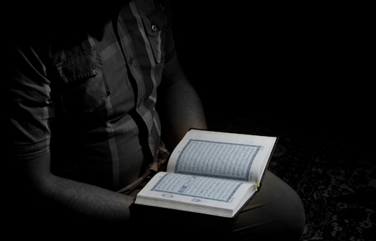 A person holding an open book, likely a religious text, illuminated by a focused light in an otherwise dark setting. The person is partially visible, wearing a checkered shirt.