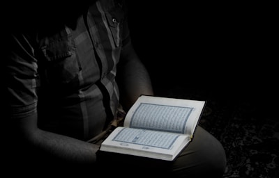 A person holding an open book, likely a religious text, illuminated by a focused light in an otherwise dark setting. The person is partially visible, wearing a checkered shirt.