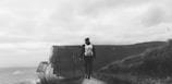A solitary person wearing a backpack is walking along a narrow path that winds atop a cliff. The landscape is vast and open, with the sea visible in the distance under a cloudy sky. The scene conveys a sense of isolation and exploration.