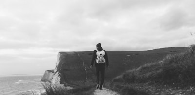 A solitary person wearing a backpack is walking along a narrow path that winds atop a cliff. The landscape is vast and open, with the sea visible in the distance under a cloudy sky. The scene conveys a sense of isolation and exploration.
