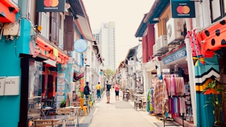 A vibrant street scene with colorful shop houses lining both sides. The shops display various items, including textiles and artwork. People are casually walking down the street, and some are sitting at small outdoor tables. The architecture is a mix of traditional and modern styles.
