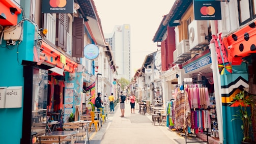 A vibrant street scene with colorful shop houses lining both sides. The shops display various items, including textiles and artwork. People are casually walking down the street, and some are sitting at small outdoor tables. The architecture is a mix of traditional and modern styles.