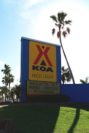 A large sign with a yellow background and red logo reads 'KOA Holiday' and lists amenities such as a pool, hot tub, Wi-Fi, propane, sunset deck, and pet playground. The sign is situated on a grassy area with several palm trees around, and the sunlight is visibly shining through the leaves of one of the trees.
