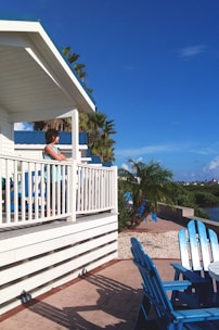 A person stands on a white wooden deck that overlooks a waterside view. The deck is part of a building with a white and blue color scheme. Palm trees and vibrant blue skies enhance the tropical setting, with blue wooden chairs scattered on a tiled patio below the deck.