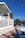 A person stands on a white wooden deck that overlooks a waterside view. The deck is part of a building with a white and blue color scheme. Palm trees and vibrant blue skies enhance the tropical setting, with blue wooden chairs scattered on a tiled patio below the deck.