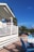 A person stands on a white wooden deck that overlooks a waterside view. The deck is part of a building with a white and blue color scheme. Palm trees and vibrant blue skies enhance the tropical setting, with blue wooden chairs scattered on a tiled patio below the deck.