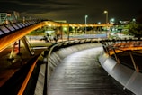 Curved walkway with sleek metal and wooden design, illuminated by artificial streetlights and architectural lighting. Modern urban environment with intersecting pathways and light trails from passing vehicles, set against a night backdrop.