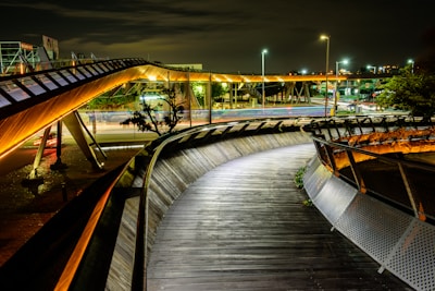 Curved walkway with sleek metal and wooden design, illuminated by artificial streetlights and architectural lighting. Modern urban environment with intersecting pathways and light trails from passing vehicles, set against a night backdrop.