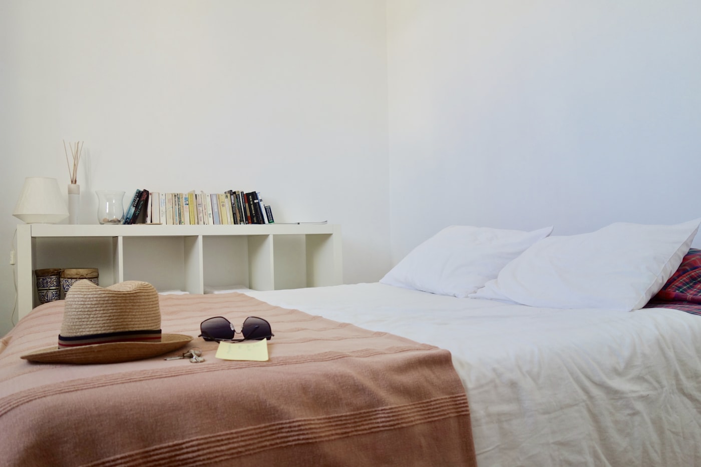 Simple apartment bedroom with white linen bedding, dusty terracotta throw blanket, and low bookshelf