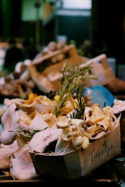 A wooden crate filled with an assortment of fresh mushrooms, including oyster and chanterelle varieties, is closely packed with sprigs of fresh rosemary. The crate is labeled with the word 'Turnips' and the background is blurred, giving the scene a rustic market feel.