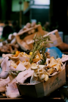 A wooden crate filled with an assortment of fresh mushrooms, including oyster and chanterelle varieties, is closely packed with sprigs of fresh rosemary. The crate is labeled with the word 'Turnips' and the background is blurred, giving the scene a rustic market feel.