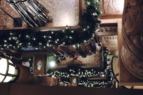 A detailed view of a decorated interior with garlands of green foliage and small white lights draped over the banisters. Marble and intricate woodwork add to the elegant architectural setting. The warm ambient lighting creates a festive and inviting atmosphere.