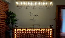 A warmly lit reception area with a modern, minimalist design. An illuminated circular chandelier hangs above a front desk with a decorative lattice pattern. A plant in the corner adds a touch of greenery beside brick-style wall accents. The words 'Maati by sundays the spa' are displayed on the wall.